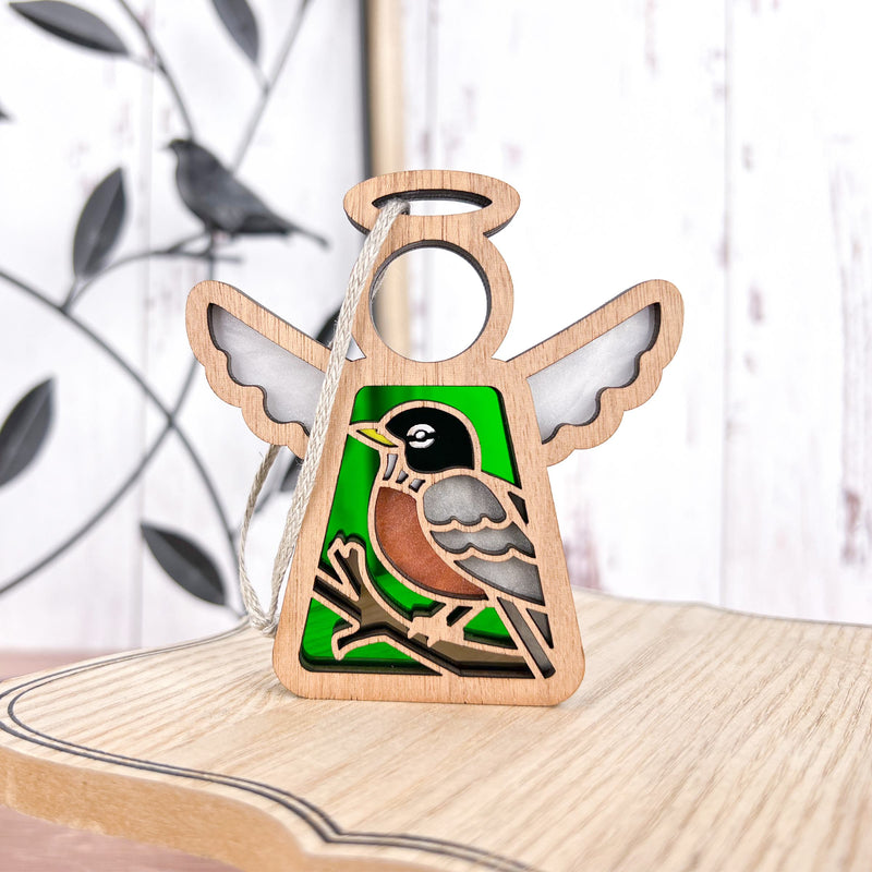 The American Robin Ornament by Forged Flare® is a 3.5" wooden Mother's Angels® figurine featuring a painted robin on a green background—perfect for bird lovers. A blurred metal bird wall decor appears in the distance.