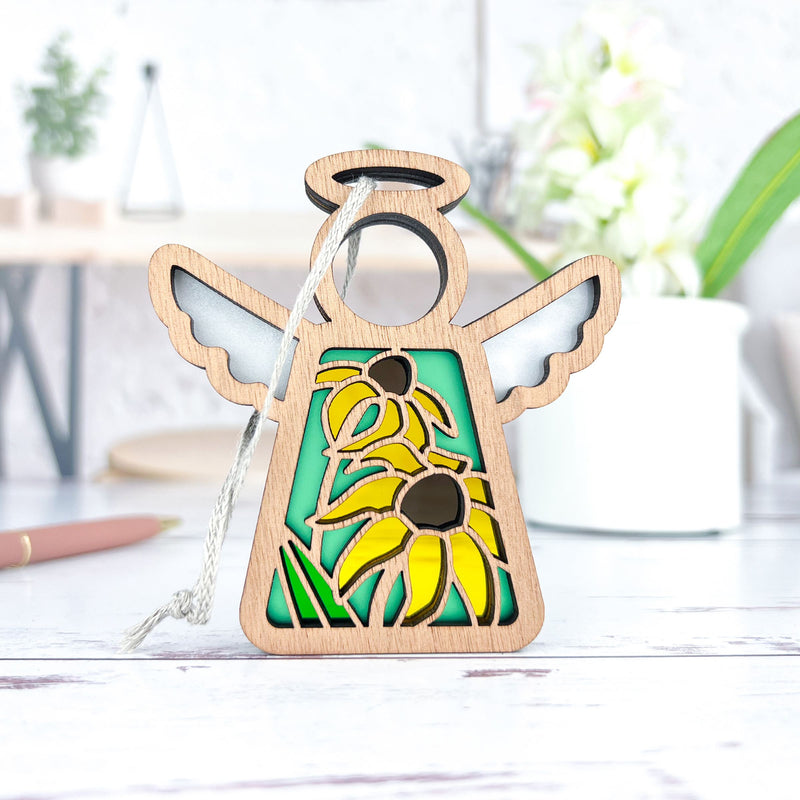 The Forged Flare® Mother's Angels® 3.5" Black-Eyed Susan Ornament features a wooden angel with stained glass-style wings and three Black-eyed Susans, displayed on a white surface with blurred plants and objects in the background.