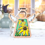 The Forged Flare® Black-Eyed Susan Ornament is a 3.5" Mother's Angels® figurine with white wings, a halo, brown-centered yellow flowers on green, and a hanging string. The background features blurred vases in autumn colors.