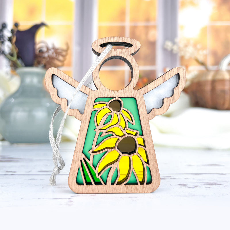 The Forged Flare® Black-Eyed Susan Ornament is a 3.5" Mother's Angels® figurine with white wings, a halo, brown-centered yellow flowers on green, and a hanging string. The background features blurred vases in autumn colors.