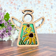 The Forged Flare® Black-Eyed Susan Ornament, a 3.5" wooden Mother's Angels® figurine with yellow flowers and green leaves cut out, stands on wood with purple and white blossoms in the background.