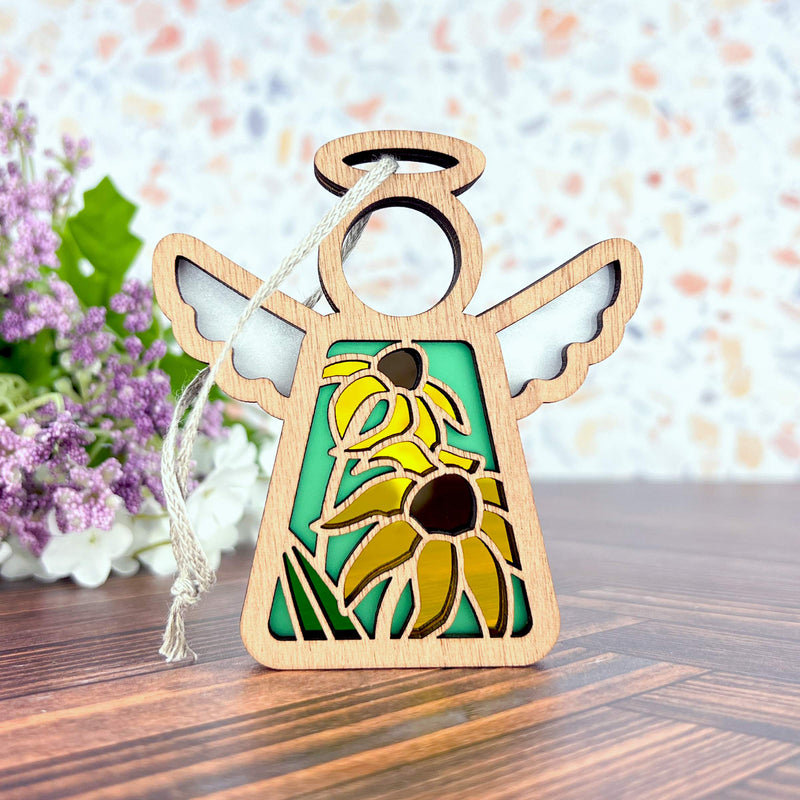The Forged Flare® Black-Eyed Susan Ornament, a 3.5" wooden Mother's Angels® figurine with yellow flowers and green leaves cut out, stands on wood with purple and white blossoms in the background.