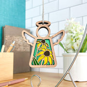 The Forged Flare® Black-Eyed Susan Ornament is a 3.5" angel figurine from Mother's Angels®, featuring a cutout Black-eyed Susans design on green and hanging by a string, with pens, books, and a white flower in the background.