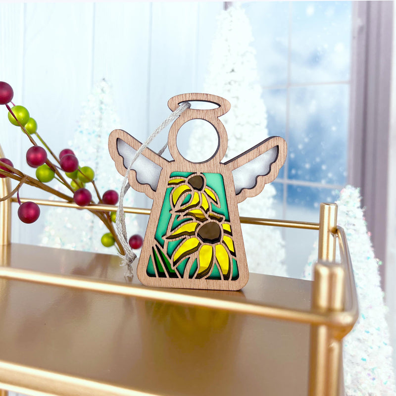 A Forged Flare® Black-Eyed Susan Ornament, a 3.5" Mother's Angels® wooden angel figurine, featuring yellow blossoms and red-green berries, sits on a gold shelf with blurred snow-covered trees and a wintry window in the background.