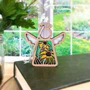 The Forged Flare® Black-Eyed Susan Ornament, a 3.5" Mother's Angels® wooden angel figurine, is perched on a green book with a garden and leafy plant visible through a large window behind it.