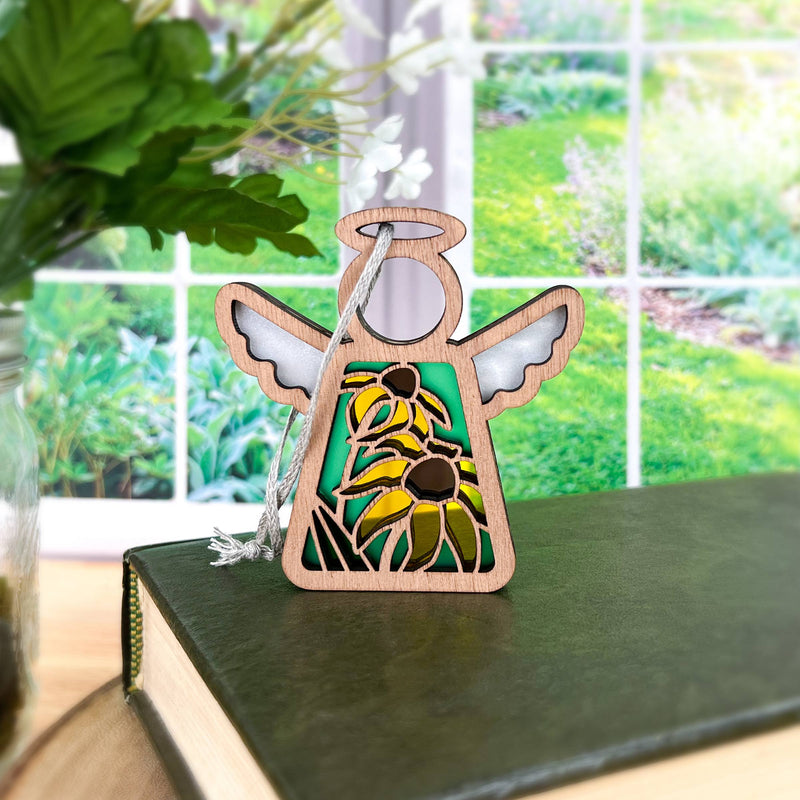 The Forged Flare® Black-Eyed Susan Ornament, a 3.5" Mother's Angels® wooden angel figurine, is perched on a green book with a garden and leafy plant visible through a large window behind it.