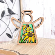 The Forged Flare® Black-Eyed Susan Ornament is a 3.5" wooden Mother’s Angels® angel figurine with metal wings and cut-out Black-eyed Susan flowers, shown standing on wood with a blurred black bird Christmas ornament behind.
