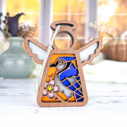 Forged Flare® Mother's Angels® 3.5" Blue Jay Ornament—an angel figurine with a stained-glass-look blue jay and pink flower—stands upright on a white surface, making an ideal gift for bird lovers. Soft-focus vases and a window appear in the background.