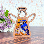 The Forged Flare® Blue Jay Ornament is a 3.5" Mother's Angels® figurine, featuring stained glass details with a blue jay and pink flower—a thoughtful gift for bird lovers. It stands on wood with purple flowers in the background.