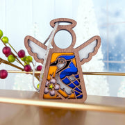 The Forged Flare® Blue Jay Ornament | 3.5" (H) Mother's Angels® figurine features a charming angel design, hanging from a silver ribbon—an ideal gift for bird lovers, beautifully set with frosted trees and berries in the background.