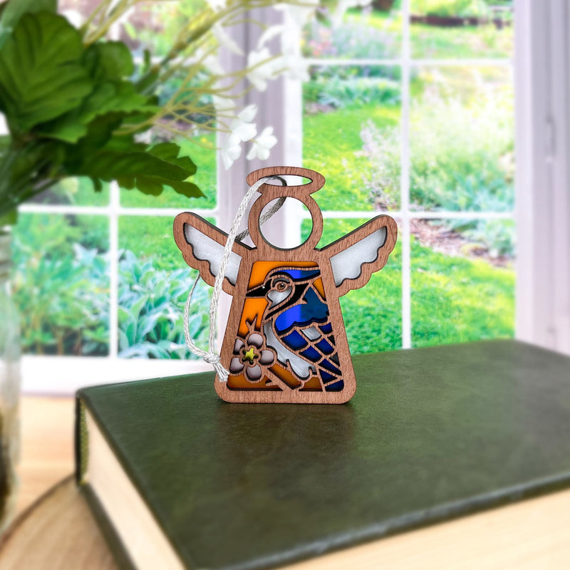 The Forged Flare® Mother's Angels® Blue Jay Ornament, a 3.5" stained glass angel figurine with bird and flower design, rests on a green book with garden views in the background—an ideal gift for bird lovers.