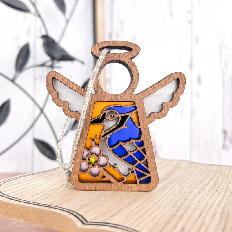 The Forged Flare® Blue Jay Ornament | 3.5" (H) Mother's Angels® angel figurine features a stained glass-style blue bird and pink flower—an ideal gift for bird lovers. A blurred black bird decor appears in the background.