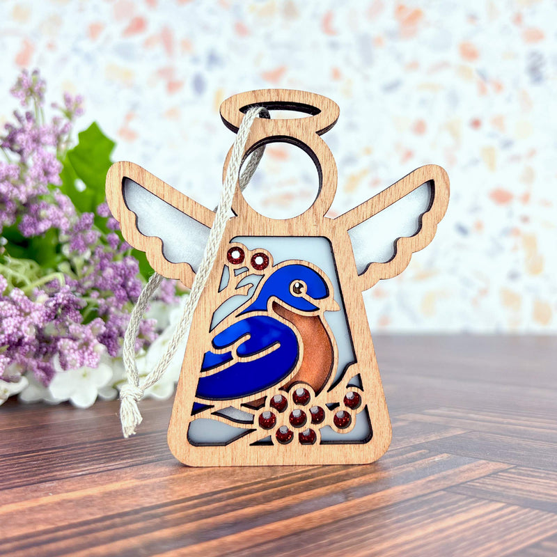 The Bluebird Ornament by Forged Flare® is a 3.5" Mother's Angels® angel figurine, featuring a bluebird and red berries with a silver hanging string. Ideal as a memorial or bird lover gift, it’s shown on wood with purple flowers behind.