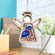 The Forged Flare® Bluebird Ornament, a 3.5" Mother's Angels® figurine, hangs indoors near a mirror with a blurred desk and potted flowers, creating a charming Bird Lover Gift with its colorful design.