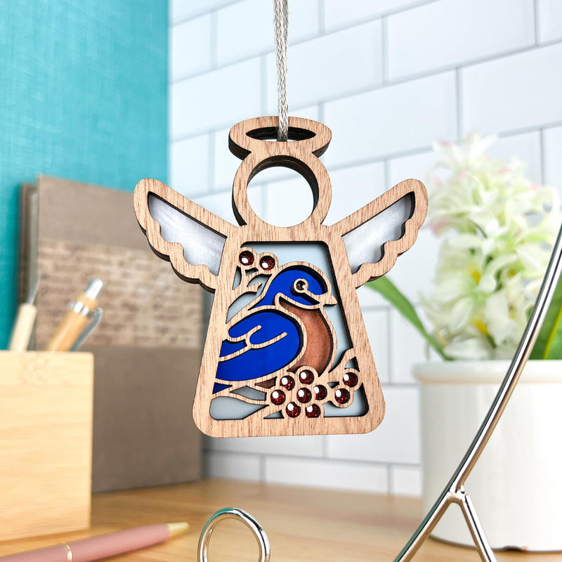 The Forged Flare® Bluebird Ornament, a 3.5" Mother's Angels® figurine, hangs indoors near a mirror with a blurred desk and potted flowers, creating a charming Bird Lover Gift with its colorful design.