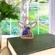 The Bluebird Ornament | 3.5" (H) Angel Figurine by Forged Flare® from Mother's Angels® rests on a green book, featuring a painted bluebird and flowers—a charming bird lover gift with garden vibes.