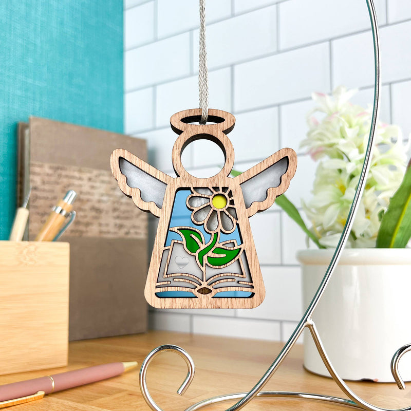 The Forged Flare® Book Lovers Ornament | 3.5" (H) Angel Figurine from Mother's Angels® features cut-out wings, a yellow flower, and a book design—perfect as a book club or author gift and displayed indoors with books and flowers.
