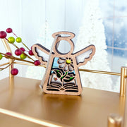The Book Lovers Ornament by Forged Flare® is a 3.5" Mother's Angels® figurine, featuring an angel with sunflower and greenery on a book, standing on a gold shelf—ideal as an author gift or for book club members. White trees and berry décor complete the festive scene.