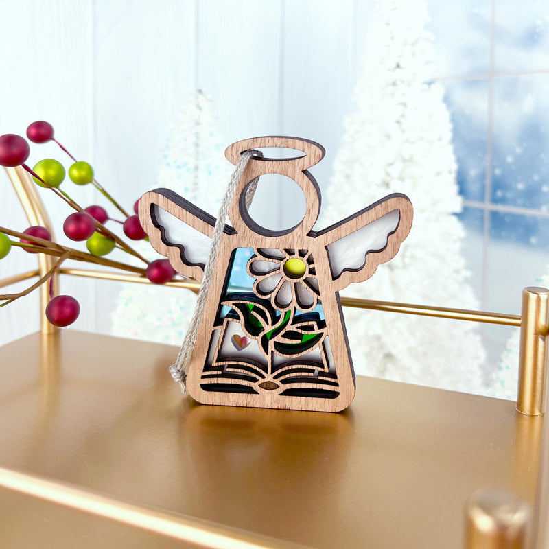 The Book Lovers Ornament by Forged Flare® is a 3.5" Mother's Angels® figurine, featuring an angel with sunflower and greenery on a book, standing on a gold shelf—ideal as an author gift or for book club members. White trees and berry décor complete the festive scene.