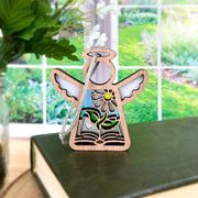 The Forged Flare® Book Lovers Ornament features a 3.5" Mother's Angels® wooden angel with floral and book details, standing on a green book near a window with greenery—an ideal gift for book clubs or authors.