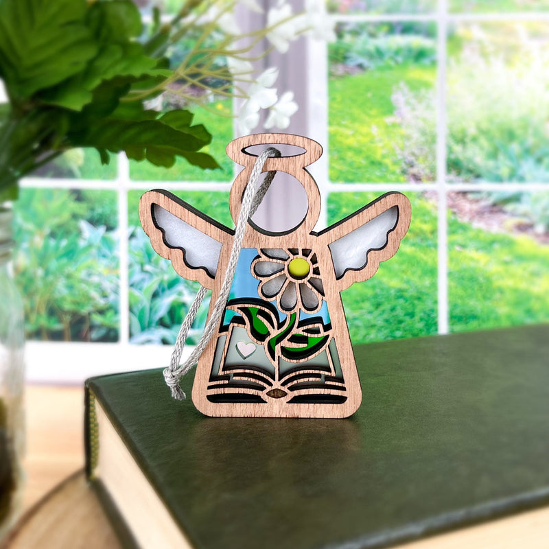 The Forged Flare® Book Lovers Ornament features a 3.5" Mother's Angels® wooden angel with floral and book details, standing on a green book near a window with greenery—an ideal gift for book clubs or authors.