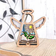 The Book Lovers Ornament by Forged Flare® is a 3.5" Mother's Angels® angel figurine with a yellow flower, green leaves, and a heart-topped open book—a charming gift for book clubs, set against metal wall art and a white wooden wall.