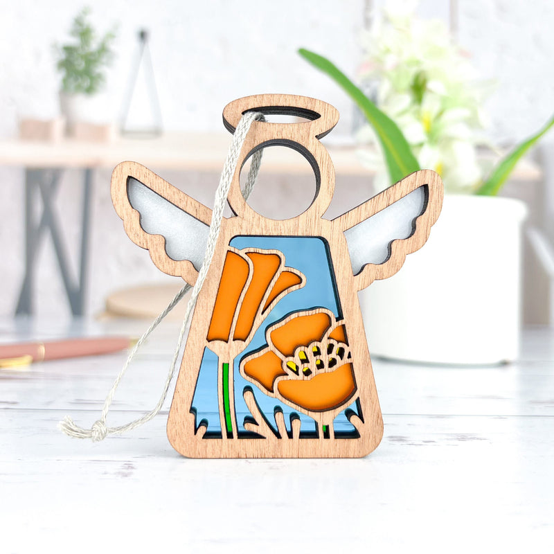 The Forged Flare® California Poppy Ornament is a 3.5" Mother's Angels® wooden angel figurine with white wings, blue accents, and orange poppy flowers, displayed with a hanging string as a charming Christmas tree ornament.