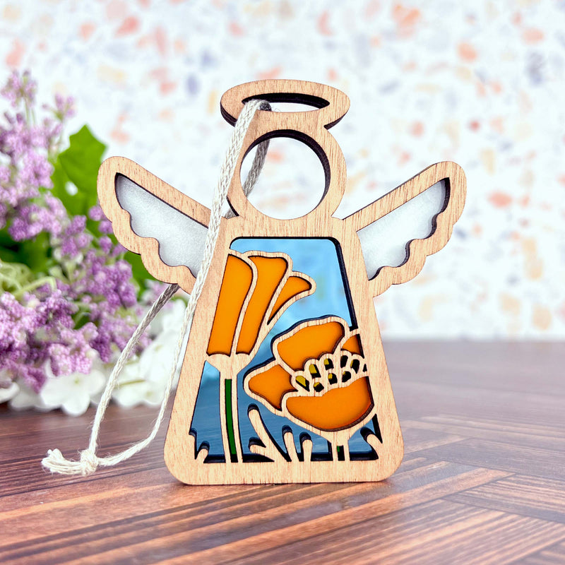 The California Poppy Ornament by Forged Flare® is a 3.5" angel figurine from the Mother's Angels® collection, featuring delicate wooden cut-outs adorned with vibrant California poppies on its body. The angel boasts simple white wings and a halo at the top, set against a background of blurred purple and white blossoms. This exquisite piece makes for a lovely Christmas ornament.