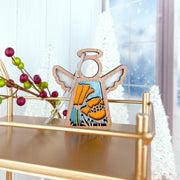 The Forged Flare® California Poppy Ornament is a 3.5" Mother's Angels® angel figurine, featuring yellow flowers, displayed on a gold shelf with frosted trees and festive red and green berries in the background.