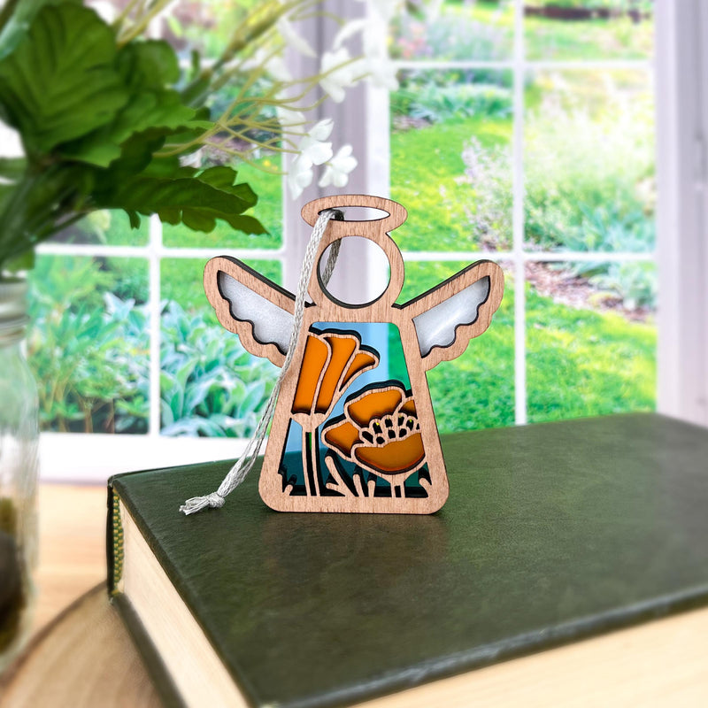 The Forged Flare® California Poppy Ornament, a 3.5" Mother's Angels® figurine with orange floral designs, stands on a green book beside a vase of leaves, with a bright garden visible through the window behind.