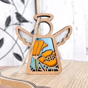 The California Poppy Ornament by Forged Flare® is a 3.5" wooden angel figurine from the Mother's Angels® line, featuring yellow poppies and green stems on a white-blue background with a string for hanging. Blurred decor sits behind it.
