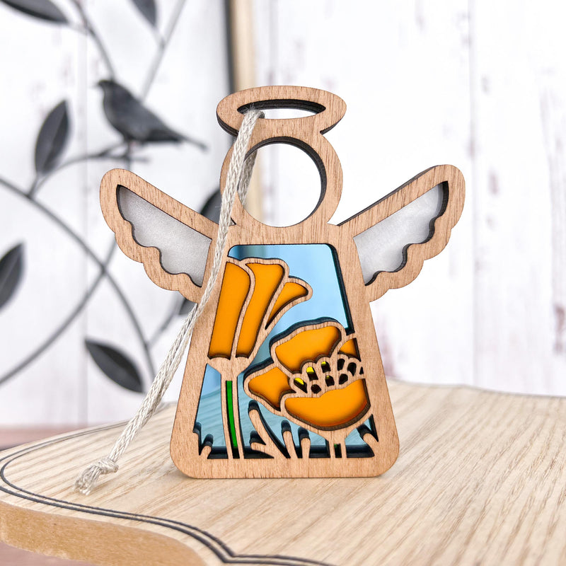 The California Poppy Ornament by Forged Flare® is a 3.5" wooden angel figurine from the Mother's Angels® line, featuring yellow poppies and green stems on a white-blue background with a string for hanging. Blurred decor sits behind it.