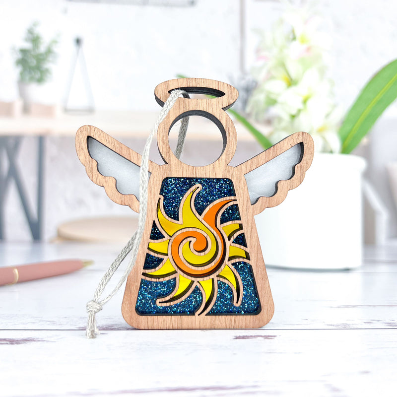 The Forged Flare® Celestial Sun Ornament, a 3.5" Mother's Angels® wooden angel figurine with a yellow and orange sun on a dark blue glittery background, has a white cord for hanging and is shown on a white surface with blurred indoor plants.