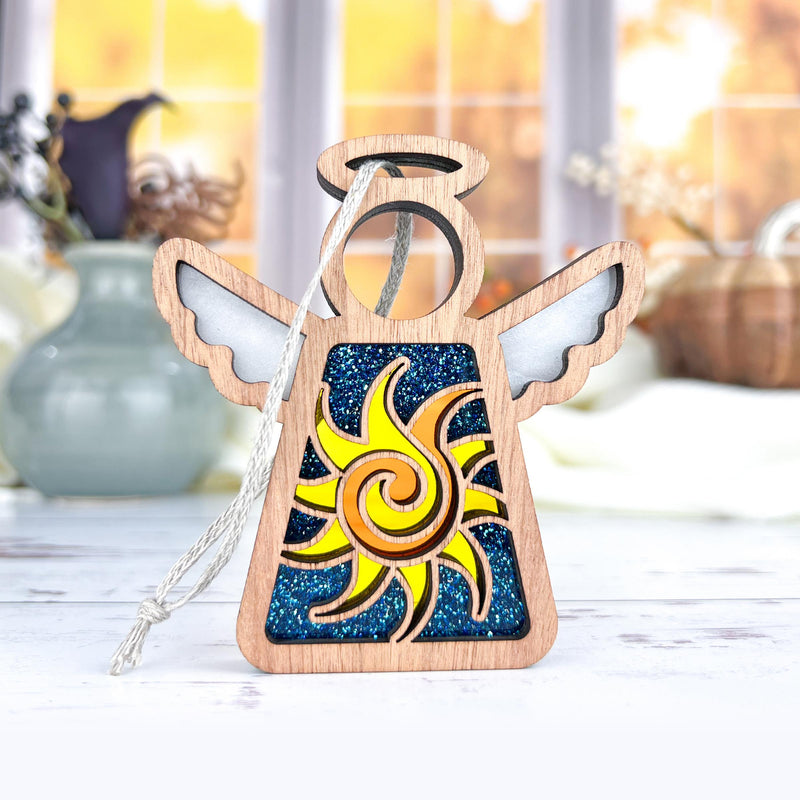 The Celestial Sun Ornament by Forged Flare® is a 3.5" wooden Mother's Angels® figurine with white wings, a halo, and a yellow sun on blue glitter—ideal as a Christmas ornament or beach-themed gift. Displayed with autumn accents in the background.