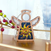 The Forged Flare® Celestial Sun Ornament features a 3.5" Mother's Angels® wooden angel with a sun design and glittery blue backdrop, hanging by a string amid festive berries and frosted trees for a charming Christmas display.