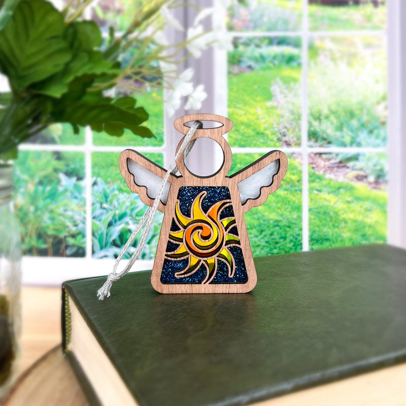 The Celestial Sun Ornament by Forged Flare® is a 3.5" Mother's Angels® figurine featuring a yellow sun and dark blue background, standing on a green book with garden views—an enchanting Christmas ornament for any space.
