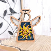 The Celestial Sun Ornament by Forged Flare® is a 3.5" Mother's Angels® angel figurine with cut-out wings and a yellow sun on a blue glittery body, shown on wood with a black bird—a charming Christmas or beach gift.
