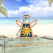 The Celestial Sun Ornament, a 3.5" angel figurine from Forged Flare® Mother's Angels®, stands on a weathered turquoise surface with seashells, palm trees, and blue ocean—making it a perfect beach-themed Christmas gift.
