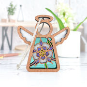 The Forged Flare® Columbine Ornament is a 3.5" Mother's Angels® figurine featuring stained glass effect wings and a blue-purple floral design inspired by the Columbine flower, displayed on a white surface with plants in the background.