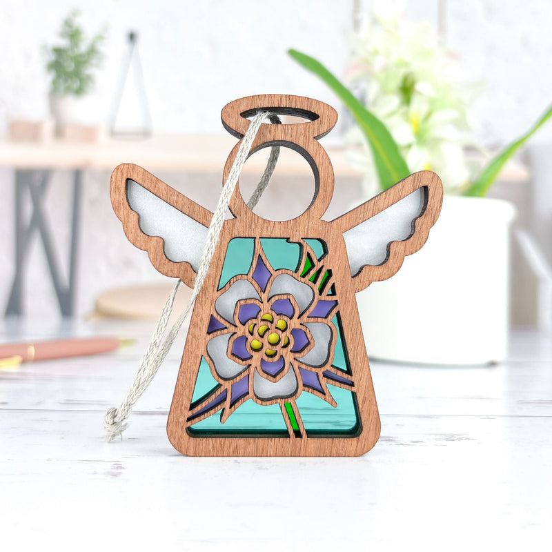 The Forged Flare® Columbine Ornament is a 3.5" Mother's Angels® figurine featuring stained glass effect wings and a blue-purple floral design inspired by the Columbine flower, displayed on a white surface with plants in the background.