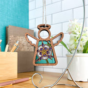 The Columbine Ornament, a 3.5" angel figurine from Forged Flare® Mother's Angels®, features a floral design and turquoise background, hanging on a metal stand among pens, books, and a potted plant against white tile.