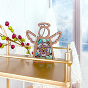 The Forged Flare® Columbine Ornament is a 3.5" Mother's Angels® figurine featuring colorful flowers, red and green berries on the left, and stands on a gold shelf with snowy trees and a frosted window in the background.