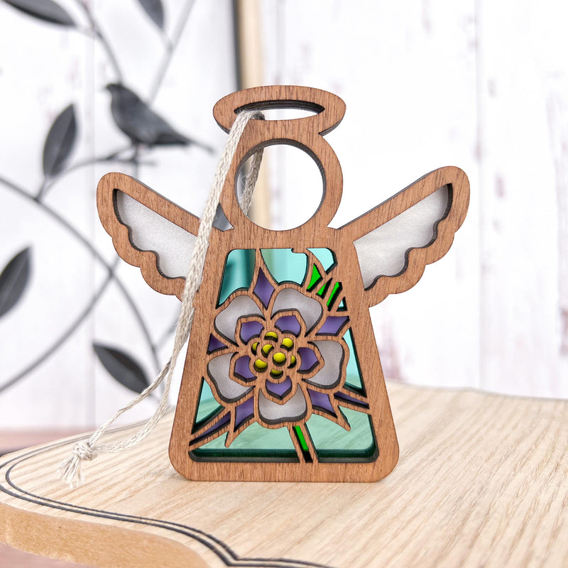 The Forged Flare® Columbine Ornament is a 3.5" Mother's Angels® wooden angel figurine with a floral stained glass center, purple petals, green accents, and an attached string for hanging as a lovely Christmas tree decoration.