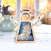 The Forged Flare® Mother's Angels® Feather Ornament is a 3.5" wooden angel figurine with white wings, halo, and blue feather inset—ideal as a Christmas or mindfulness gift. Shown standing on a light surface near vases with autumn decor.