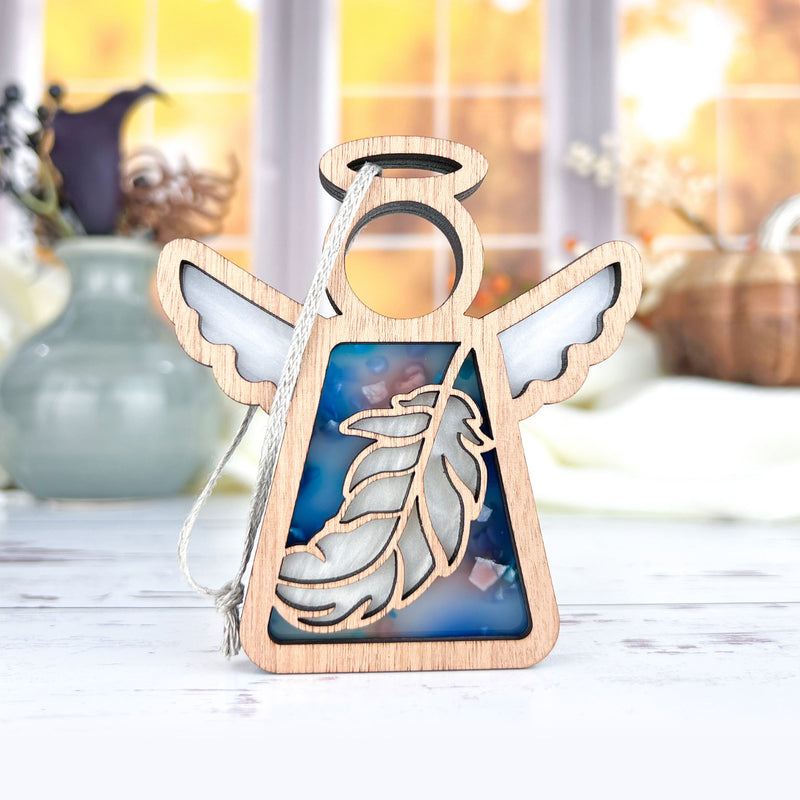 The Forged Flare® Mother's Angels® Feather Ornament is a 3.5" wooden angel figurine with white wings, halo, and blue feather inset—ideal as a Christmas or mindfulness gift. Shown standing on a light surface near vases with autumn decor.