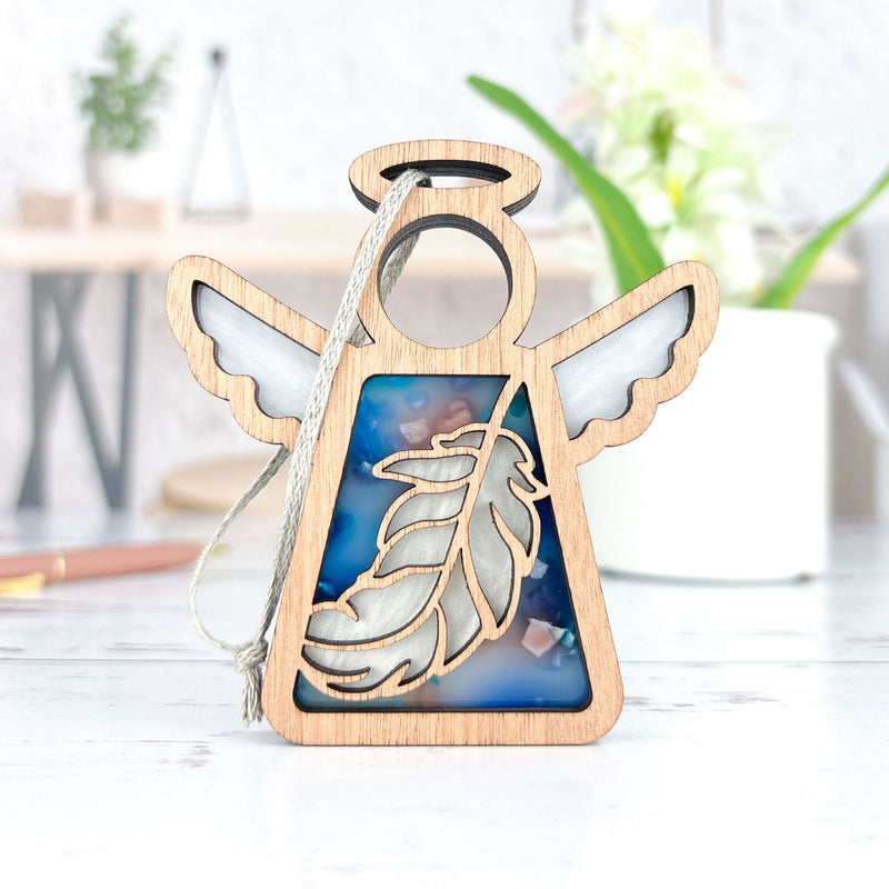 The Feather Ornament | 3.5" (H) Angel Figurine by Forged Flare® from Mother's Angels® is a wooden angel with blue and white resin feather details—a perfect Christmas ornament or mindfulness gift, elegantly displayed with blurred decor in the background.
