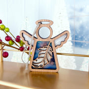 The Forged Flare® Mother's Angels® Feather Ornament is a 3.5" wooden angel figurine with translucent blue accents, hanging by a white string—an ideal Christmas ornament or mindfulness gift. Red berries and white trees are softly blurred in the background.