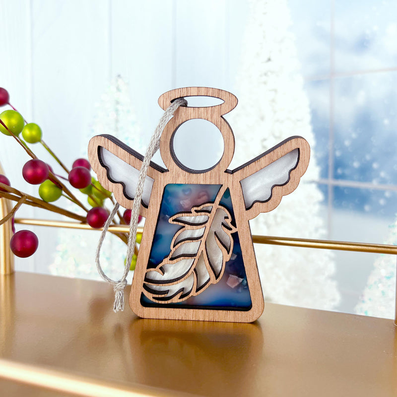 The Forged Flare® Mother's Angels® Feather Ornament is a 3.5" wooden angel figurine with translucent blue accents, hanging by a white string—an ideal Christmas ornament or mindfulness gift. Red berries and white trees are softly blurred in the background.