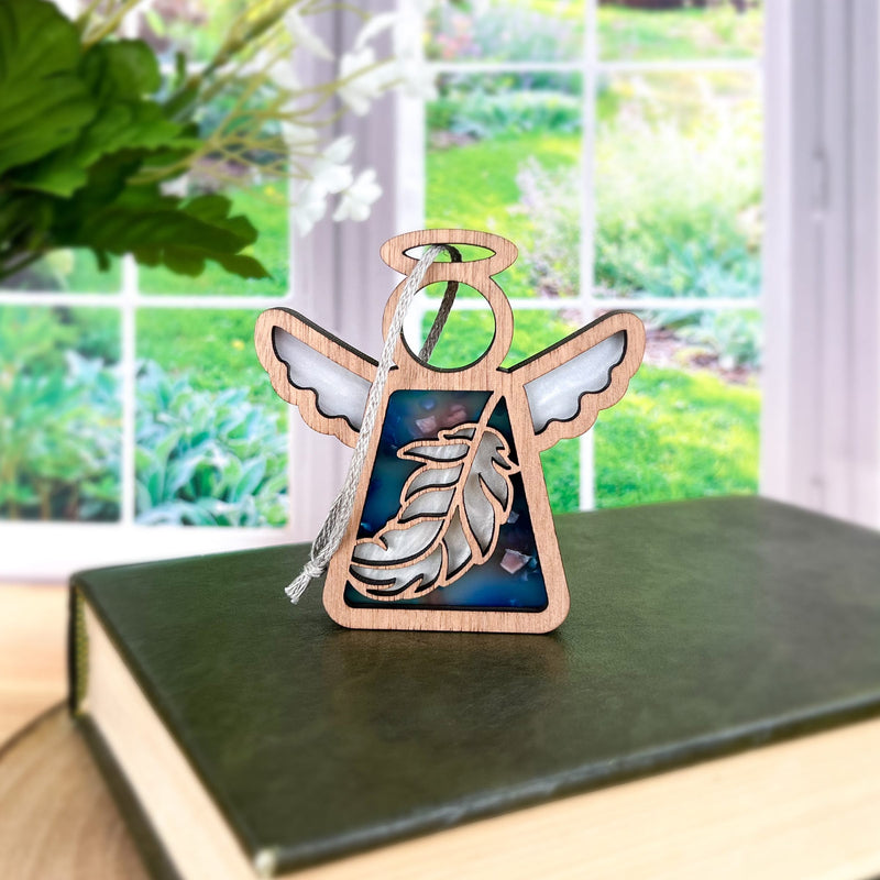 The Feather Ornament | 3.5" (H) Angel Figurine by Forged Flare® sits atop a closed green book, making an ideal mindfulness gift. A bright window behind reveals a lush garden, enhancing the peaceful scene.