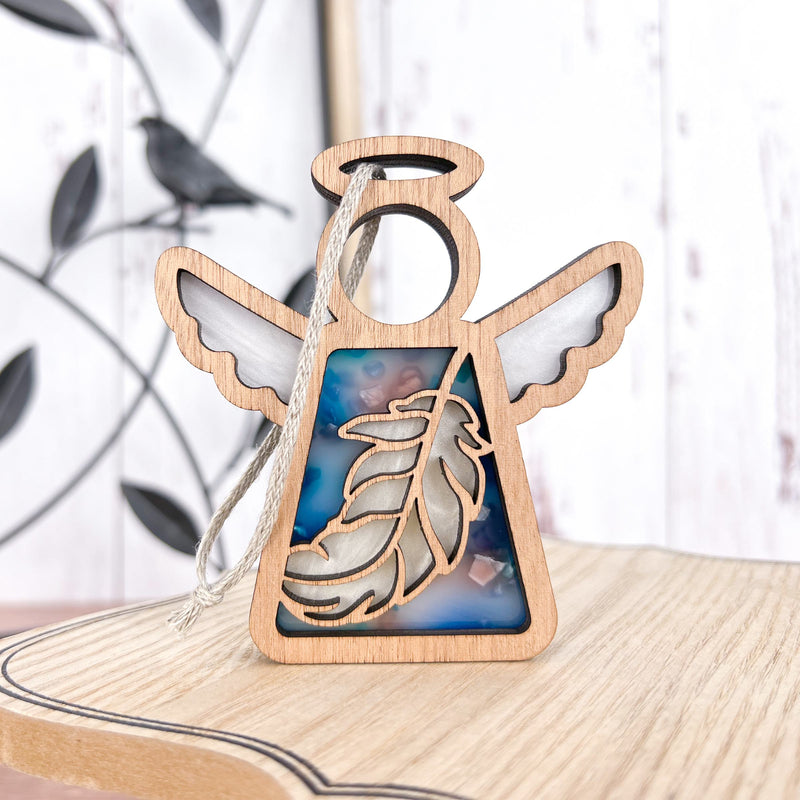 The Forged Flare® Mother's Angels® Feather Ornament is a 3.5" wooden angel figurine with white wings, a halo, and hanging string, featuring a feather design over blue at the center. It sits on light wood with blurred bird decor in the background.